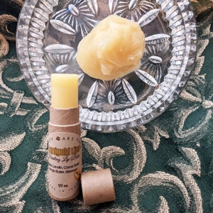 Può includere: Un tubo marrone di balsamo per labbra con il testo "Emerald Lips Healing Lip Balm" e un elenco di ingredienti tra cui "Lime, Lavanda, Burro di Cacao, Burro di Mango, Cera d'Api". Il tubo è pieno per un terzo e il balsamo per labbra è di colore giallo chiaro. Il balsamo per labbra è su un tessuto a motivi verdi e dorati accanto a un piccolo piatto con una noce di balsamo per labbra giallo.