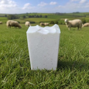 May include: A white, rectangular bar of soap with a textured surface. The soap is set in a field of green grass, with sheep grazing in the background under a blue sky.