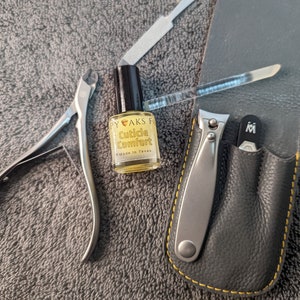 May include: A manicure set with a gray leather case, nail clippers, cuticle nippers, a nail file, and a bottle of cuticle comfort oil. The oil bottle has a black cap and yellow label with text.