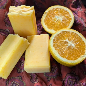 May include: Four bars of orange-scented soap and two orange halves on a red and purple patterned fabric.