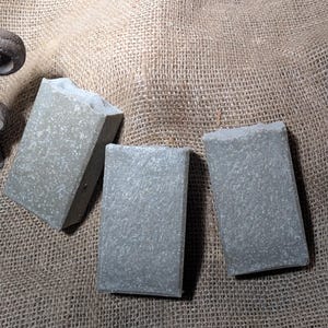 May include: Three bars of handmade soap, each with a light gray color and a speckled texture. The soap bars are arranged on a burlap surface.