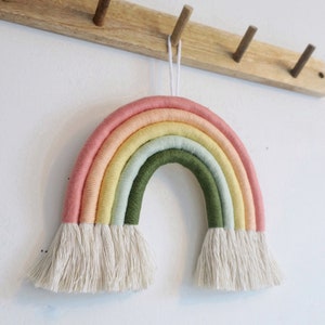 Op de afbeelding: Een handgemaakte regenboog wandhanger van macramé. De regenboog heeft lagen van roze, perzik, geel, lichtgroen en olijfgroen garen. De onderkant van de regenboog heeft een franje van crèmekleurig garen. Hangt aan een houten wandrek.