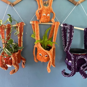 May include: Three crocheted hanging plant pots shaped like octopus with orange and purple yarn. The pots are hanging from a wooden rack on a blue wall.