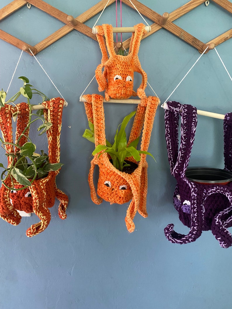 May include: Three crocheted hanging plant pots shaped like octopus with orange and purple yarn. The pots are hanging from a wooden rack on a blue wall.