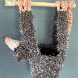 May include: A gray and pink crocheted hanging planter shaped like an opossum. The planter has a fuzzy texture and a small, white, crocheted nose with a blue eye.
