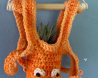 Può includere: Un vaso appeso in crochet arancione con una piccola pianta grassa al suo interno. Il vaso ha un viso con due occhi neri e un contorno bianco.