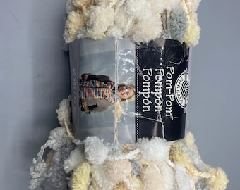 Può includere: Un gomitolo di filato bianco, grigio e giallo con pompon. Il filato è etichettato "Pom-Pom Pompon" e "All-Threads".