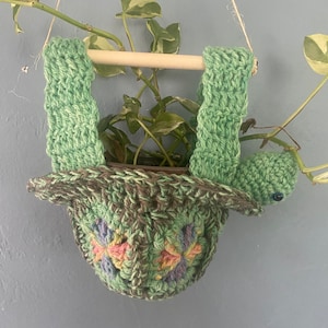 May include: A green crocheted turtle planter hanging from the ceiling with a potted plant inside. The turtle has a brown and green shell with colorful flowers crocheted into the design.