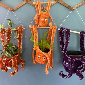 May include: Three crocheted octopus plant hangers with cute faces. The hangers are orange, orange, and purple. Each hanger has a small plant inside.