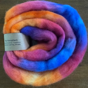 May include: A close-up of a colorful, hand-dyed roving of wool yarn in shades of orange, pink, purple, and blue. The roving is rolled into a circle and has a label that reads "The Hierophant Spiritual Guidance, Traditions, Traditional Learning, Rituals".