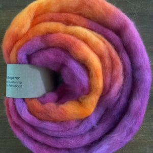 May include: A close-up of a bundle of purple and orange wool roving. The roving is arranged in a circle, with the purple wool on the outside and the orange wool on the inside. The roving is soft and fluffy, and it is perfect for spinning or felting.