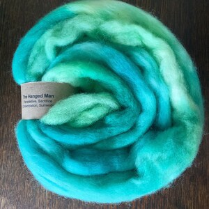 May include: A skein of turquoise, teal, and green wool yarn. The yarn is rolled into a circle and has a label that reads "The Hanged Man Perspective, Sacrifice, Contemplation, Surrender".
