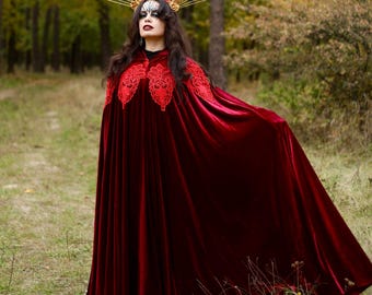 May include: A woman wearing a burgundy velvet cloak with red lace detailing on the shoulders. She is wearing a gold halo crown with a floral design and a face mask. The background is a forest with a path.