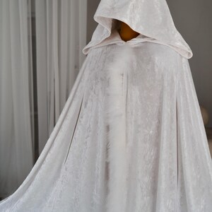 White fantasy hooded cloak made from luxurious soft velvet fabric with decorative feather trim and elegant flowing silhouette. Handmade full length cape inspired by cinematic fantasy fashion and medieval aesthetics designed for cosplay, artistic styling, renaissance inspired outfits and themed photography.