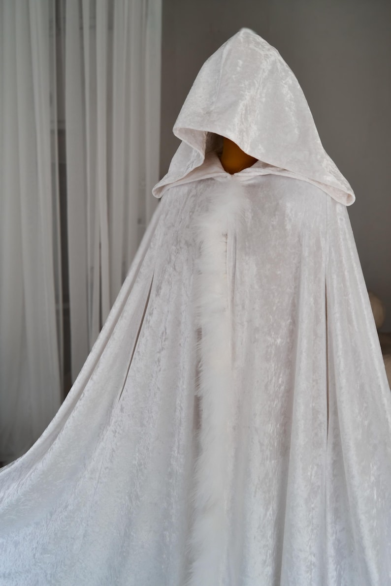 White fantasy hooded cloak made from luxurious soft velvet fabric with decorative feather trim and elegant flowing silhouette. Handmade full length cape inspired by cinematic fantasy fashion and medieval aesthetics designed for cosplay, artistic styling, renaissance inspired outfits and themed photography.