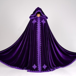 May include: A long, flowing, hooded cloak in a deep purple velvet. The cloak features intricate, lighter purple embroidered trim along the front opening, hood, and hem. The design includes floral or star-like patterns. The cloak is displayed against a plain white background.