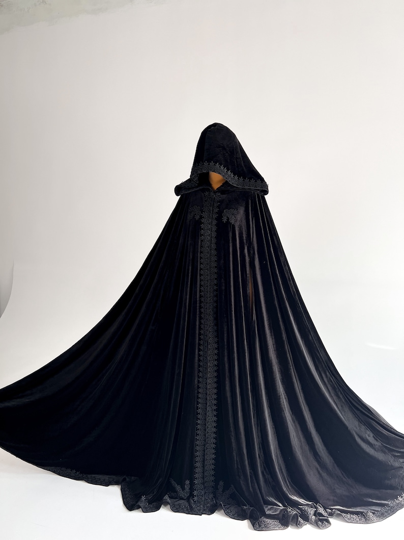 Dramatic black velvet witch cloak with oversized hood inspired by gothic Halloween fashion and fantasy cosplay aesthetics. Handmade hooded cape with elegant flowing shape and premium velvet texture designed for vampire costumes, dark feminine styling, renaissance fairs and cinematic photoshoots. Floor length silhouette creates powerful luxury gothic appearance.
