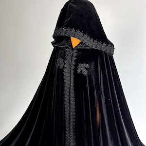 Handmade black velvet cloak with hood and hidden arm openings designed for comfortable movement during Halloween parties, gothic events and fantasy cosplay. Medium weight velvet fabric creates rich folds and luxurious texture while remaining comfortable to wear. Elegant hooded witch cape inspired by vampire aesthetics, medieval fantasy and gothic fashion trends.
