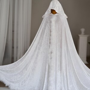 Pu&ograve; includere: Un lungo mantello bianco con cappuccio realizzato in un tessuto strutturato, forse velluto. Il mantello ha un design ampio e fluente ed &egrave; rifinito con piume bianche lungo l'orlo. L'indumento &egrave; esposto su un manichino.