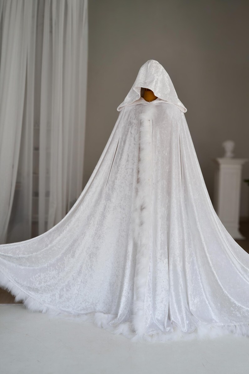 Pu&ograve; includere: Un lungo mantello bianco con cappuccio realizzato in un tessuto strutturato, forse velluto. Il mantello ha un design ampio e fluente ed &egrave; rifinito con piume bianche lungo l'orlo. L'indumento &egrave; esposto su un manichino.