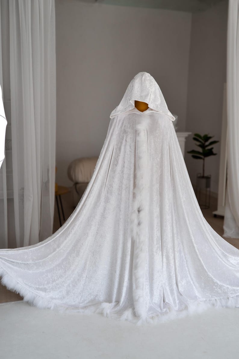 Elegant white velvet fantasy cape with oversized hood and decorative feather trim inspired by medieval and fantasy aesthetics. Handmade full length cloak with luxurious soft velvet texture and dramatic flowing silhouette perfect for cosplay, artistic photoshoots, fantasy styling and renaissance inspired fashion looks.