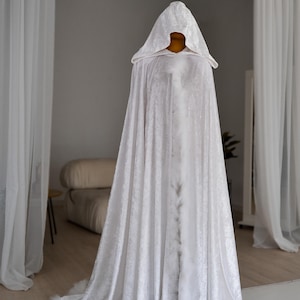 White velvet fantasy cape with oversized hood and decorative feather trim designed for elegant fantasy styling and artistic fashion looks. Handmade unisex cloak with flowing floor length silhouette and rich velvet texture perfect for cosplay, renaissance fairs, themed photography, medieval inspired outfits and fantasy aesthetics.