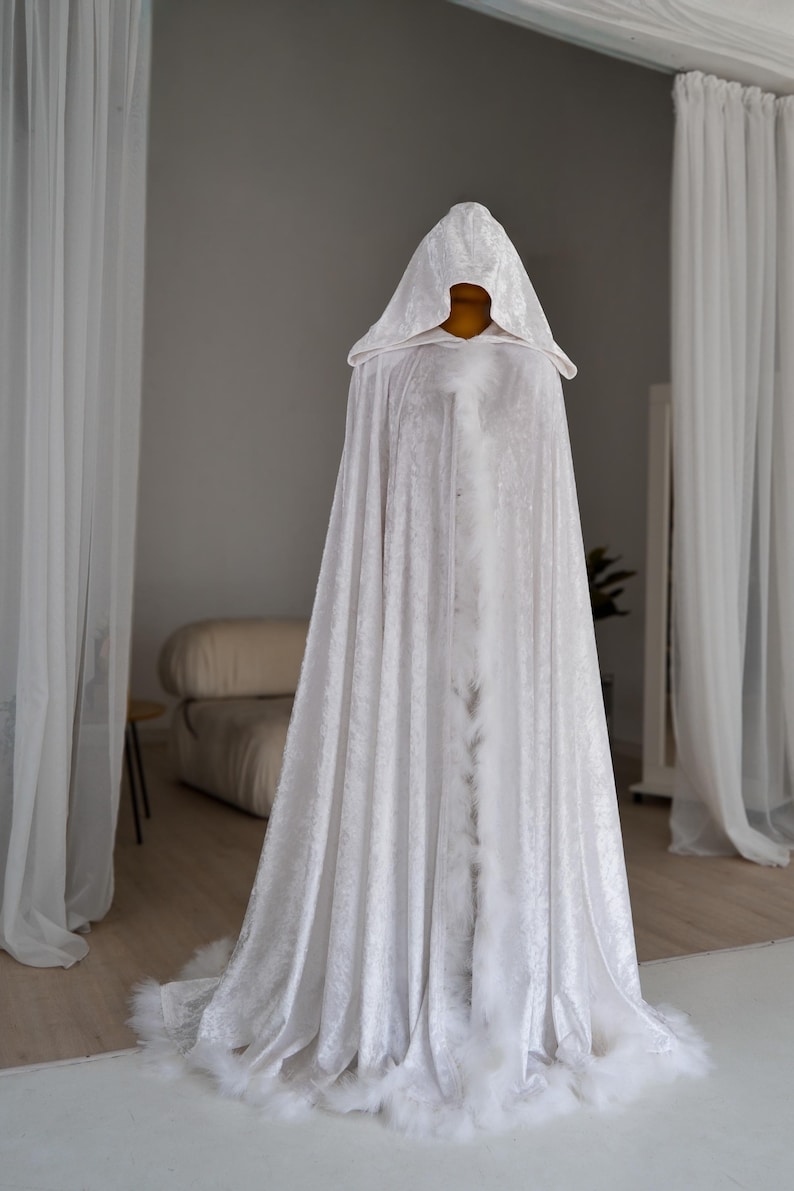 White velvet fantasy cape with oversized hood and decorative feather trim designed for elegant fantasy styling and artistic fashion looks. Handmade unisex cloak with flowing floor length silhouette and rich velvet texture perfect for cosplay, renaissance fairs, themed photography, medieval inspired outfits and fantasy aesthetics.