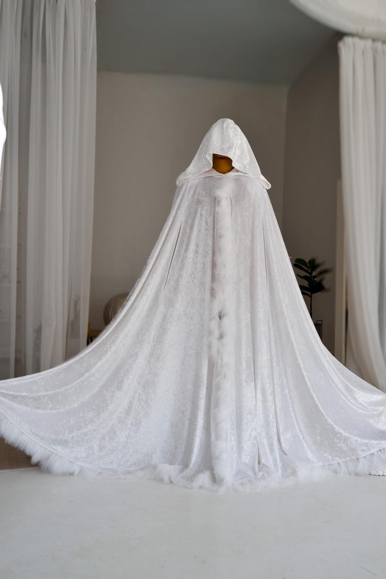 White velvet hooded cape with decorative feather trim and elegant flowing silhouette designed for fantasy aesthetics and artistic styling. Handmade unisex fantasy cloak with oversized hood and luxurious velvet texture perfect for cosplay, renaissance fairs, medieval inspired outfits and themed photoshoots.