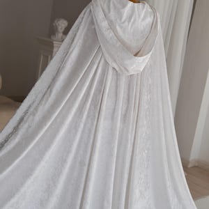 White velvet fantasy cloak with oversized hood and decorative feather trim designed for elegant fantasy inspired styling and cinematic fashion aesthetics. Handmade unisex cape with flowing silhouette and luxurious velvet texture perfect for cosplay events, artistic editorials and renaissance inspired photography.