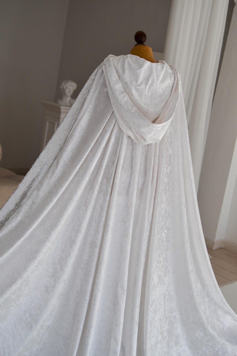 White velvet fantasy cloak with oversized hood and decorative feather trim designed for elegant fantasy inspired styling and cinematic fashion aesthetics. Handmade unisex cape with flowing silhouette and luxurious velvet texture perfect for cosplay events, artistic editorials and renaissance inspired photography.