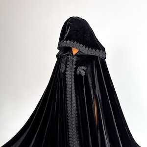 Long black velvet cloak with dramatic hood and flowing gothic silhouette inspired by dark fantasy fashion and Halloween aesthetics. Handmade hooded cape created for witch costumes, vampire cosplay, renaissance fairs and cinematic gothic styling. Medium weight premium velvet fabric creates elegant folds and luxurious movement while remaining comfortable to wear.
