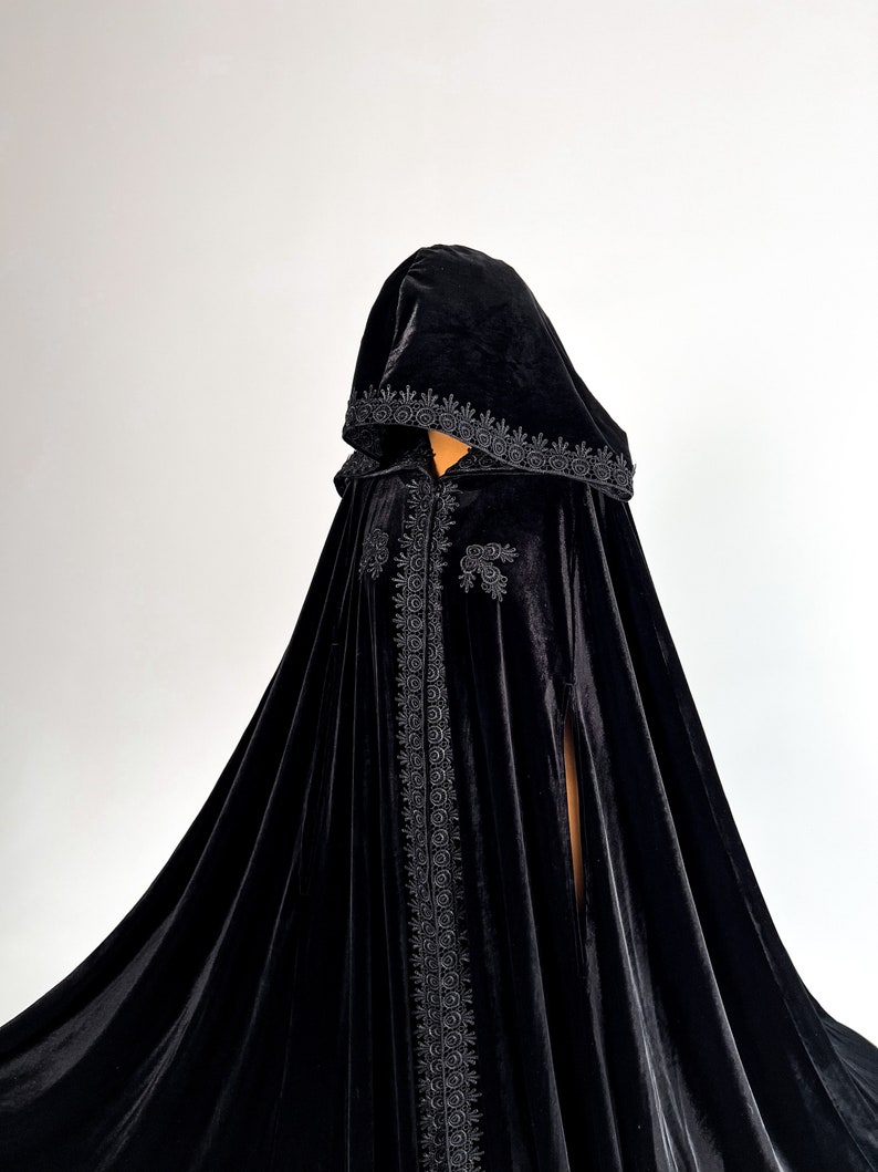 Long black velvet cloak with dramatic hood and flowing gothic silhouette inspired by dark fantasy fashion and Halloween aesthetics. Handmade hooded cape created for witch costumes, vampire cosplay, renaissance fairs and cinematic gothic styling. Medium weight premium velvet fabric creates elegant folds and luxurious movement while remaining comfortable to wear.