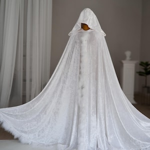 Elegant white fantasy cloak made from soft velvet fabric with luxurious texture and flowing movement. This handmade hooded cape features decorative feather trim, oversized hood and full length silhouette designed for fantasy inspired outfits, medieval aesthetics, cosplay events, renaissance fairs and artistic editorial photography.