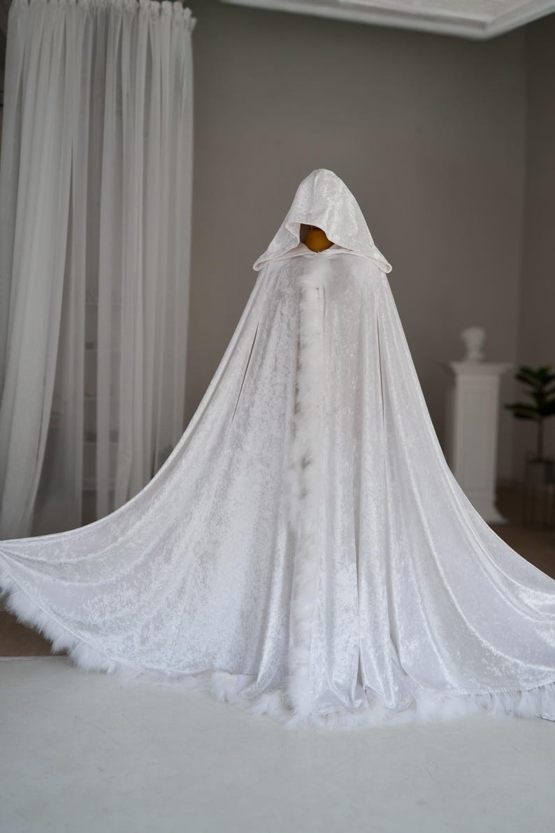 Elegant white fantasy cloak made from soft velvet fabric with luxurious texture and flowing movement. This handmade hooded cape features decorative feather trim, oversized hood and full length silhouette designed for fantasy inspired outfits, medieval aesthetics, cosplay events, renaissance fairs and artistic editorial photography.