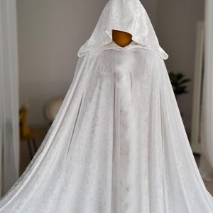 White velvet hooded fantasy cloak with decorative feather edging and elegant flowing movement. This handmade unisex cape features oversized hood, rich velvet texture and full length silhouette designed for fantasy inspired styling, cosplay photography, renaissance aesthetics, theatrical fashion and artistic editorial looks.