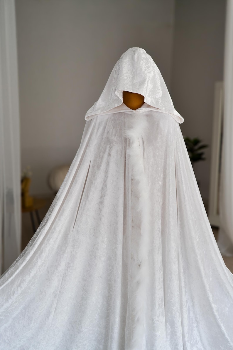 White velvet hooded fantasy cloak with decorative feather edging and elegant flowing movement. This handmade unisex cape features oversized hood, rich velvet texture and full length silhouette designed for fantasy inspired styling, cosplay photography, renaissance aesthetics, theatrical fashion and artistic editorial looks.