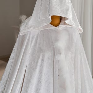 White hooded fantasy cape handmade from soft velvet fabric with luxurious texture and decorative feather trim. Elegant flowing silhouette and oversized hood create dramatic fantasy inspired appearance perfect for cosplay events, artistic editorial photography, renaissance fairs and cinematic fashion styling