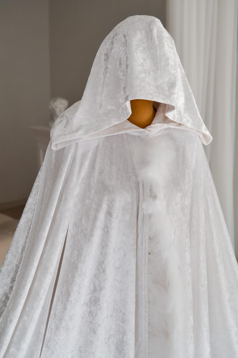 White hooded fantasy cape handmade from soft velvet fabric with luxurious texture and decorative feather trim. Elegant flowing silhouette and oversized hood create dramatic fantasy inspired appearance perfect for cosplay events, artistic editorial photography, renaissance fairs and cinematic fashion styling