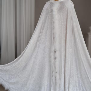 Handmade white fantasy cloak crafted from soft velvet fabric with oversized hood and decorative feather edging. This elegant full length hooded cape features flowing silhouette and luxurious texture inspired by fantasy fashion, medieval aesthetics, artistic styling and cinematic fantasy inspired photography.