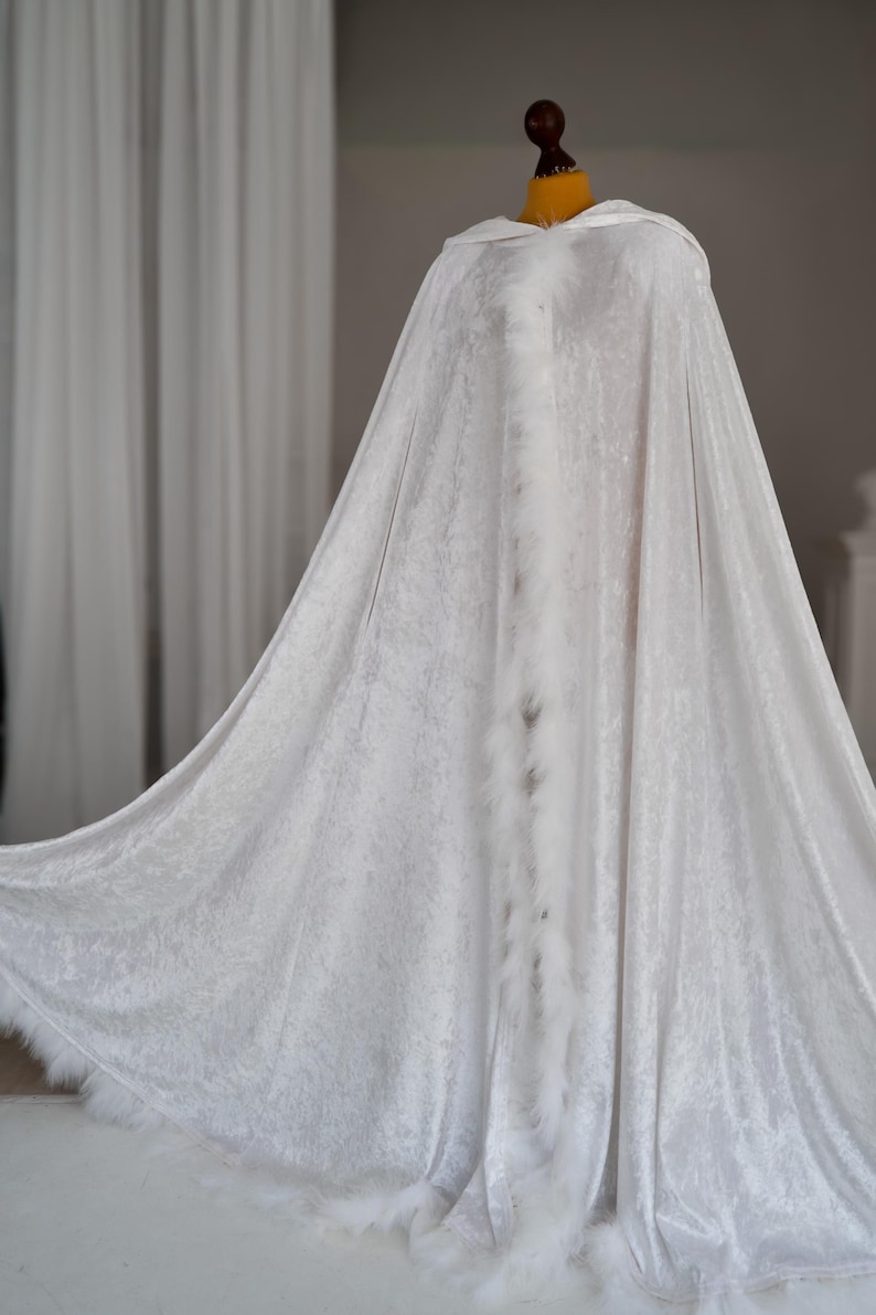 Handmade white fantasy cloak crafted from soft velvet fabric with oversized hood and decorative feather edging. This elegant full length hooded cape features flowing silhouette and luxurious texture inspired by fantasy fashion, medieval aesthetics, artistic styling and cinematic fantasy inspired photography.