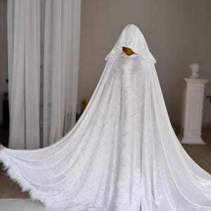 Elegant white velvet cloak with oversized hood and decorative feather edging inspired by fantasy aesthetics and medieval fashion. Handmade unisex fantasy cape with flowing floor length silhouette and rich velvet texture designed for cosplay, artistic photoshoots and renaissance inspired styling.
