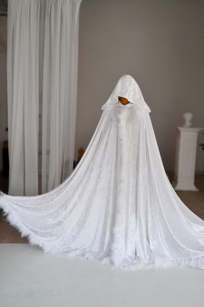Elegant white velvet cloak with oversized hood and decorative feather edging inspired by fantasy aesthetics and medieval fashion. Handmade unisex fantasy cape with flowing floor length silhouette and rich velvet texture designed for cosplay, artistic photoshoots and renaissance inspired styling.