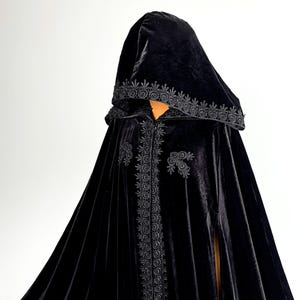 Gothic hooded cloak handmade from premium black velvet fabric with dramatic floor length silhouette and elegant movement. Halloween witch cape designed for fantasy cosplay, vampire aesthetic and renaissance inspired styling. Flowing velvet material with hidden arm slits and single button neck closure creates luxurious dark fashion appearance for themed events and photography.