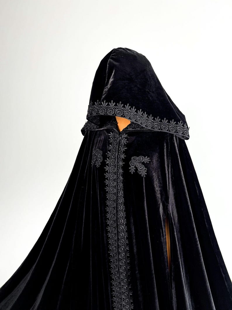 Gothic hooded cloak handmade from premium black velvet fabric with dramatic floor length silhouette and elegant movement. Halloween witch cape designed for fantasy cosplay, vampire aesthetic and renaissance inspired styling. Flowing velvet material with hidden arm slits and single button neck closure creates luxurious dark fashion appearance for themed events and photography.