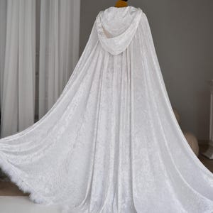 White hooded velvet cloak with flowing fantasy silhouette and decorative feather trim designed for elegant fantasy inspired outfits and cinematic styling. Handmade unisex fantasy cape with rich velvet texture, oversized hood and luxurious movement ideal for cosplay, medieval aesthetics and artistic photography.