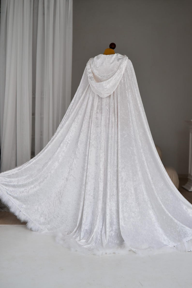 White hooded velvet cloak with flowing fantasy silhouette and decorative feather trim designed for elegant fantasy inspired outfits and cinematic styling. Handmade unisex fantasy cape with rich velvet texture, oversized hood and luxurious movement ideal for cosplay, medieval aesthetics and artistic photography.