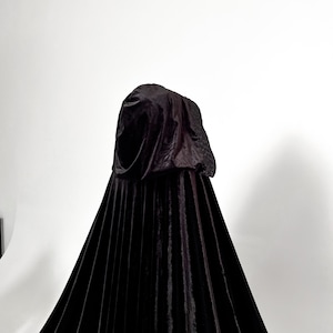 Floor length black velvet hooded cloak with dramatic gothic silhouette and elegant flowing movement. Premium handmade Halloween cape with single button neck closure and oversized hood designed for witch costumes, vampire outfits, cosplay conventions and fantasy inspired styling. Luxurious velvet texture creates cinematic dark fashion aesthetic for photoshoots and themed events.
