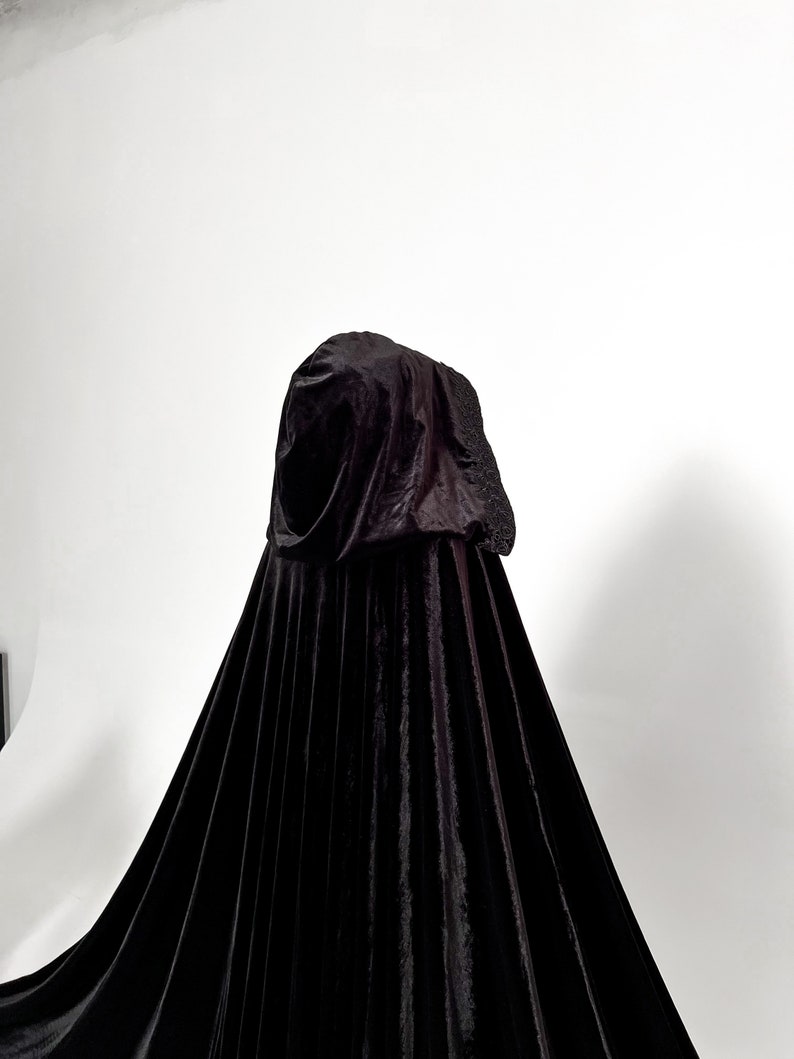 Floor length black velvet hooded cloak with dramatic gothic silhouette and elegant flowing movement. Premium handmade Halloween cape with single button neck closure and oversized hood designed for witch costumes, vampire outfits, cosplay conventions and fantasy inspired styling. Luxurious velvet texture creates cinematic dark fashion aesthetic for photoshoots and themed events.