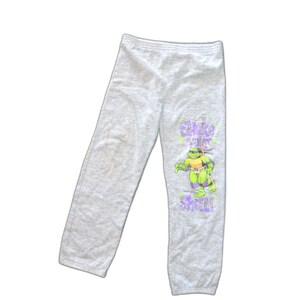 May include: Light gray sweatpants featuring a graphic of a Teenage Mutant Ninja Turtle in green, purple, and yellow. The text "Shake Your Shell!" is printed in purple and green. The pants are made of a soft material.