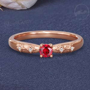 May include: Rose gold ring featuring a round, vibrant red gemstone at the center. The band is adorned with small, sparkling accent stones and decorative details. The ring is set against a dark blue background.